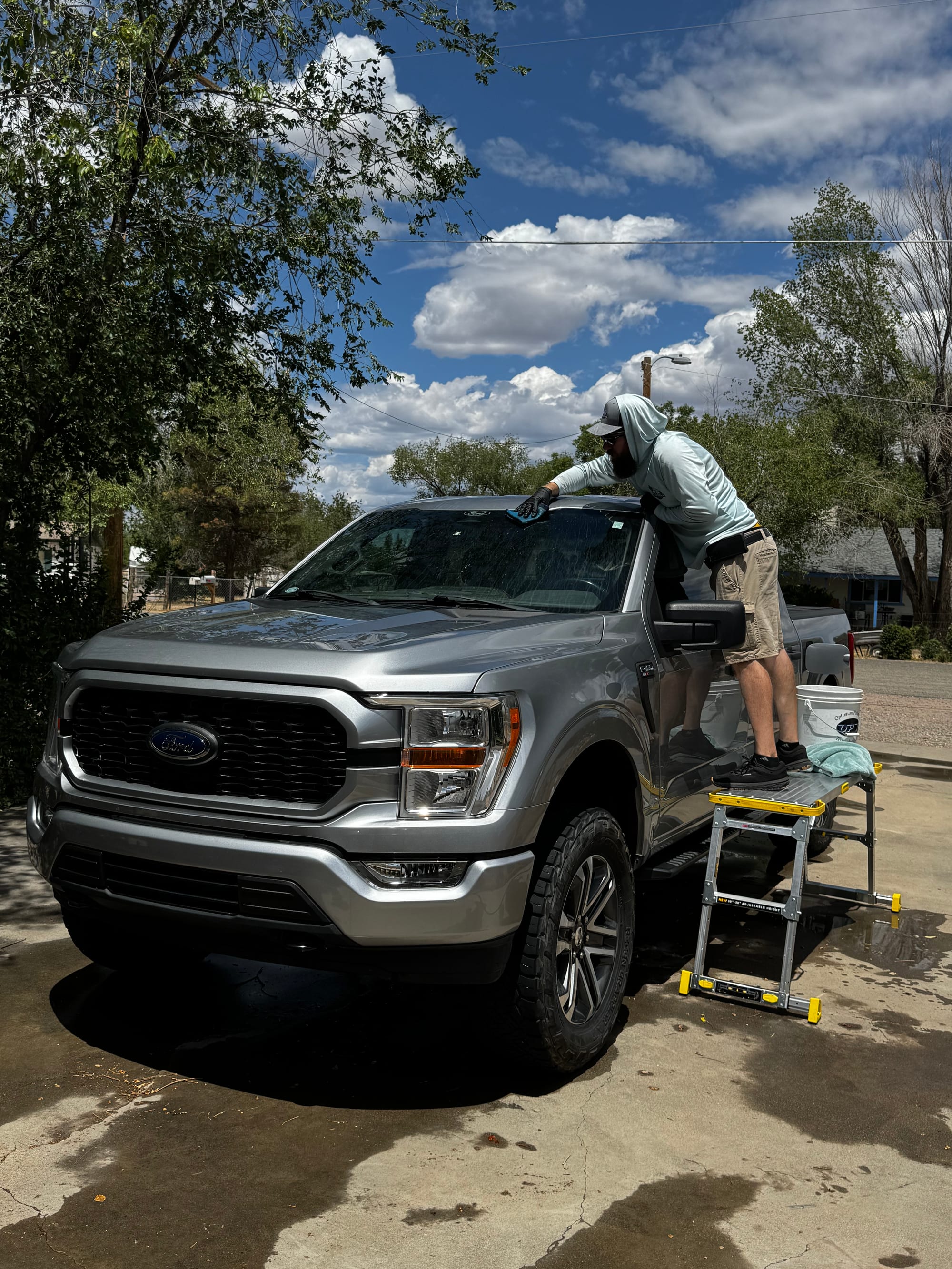 Detailer on a step-ladder hand-cleaning the roof of a Ford F-150 under a blue Arizona sky